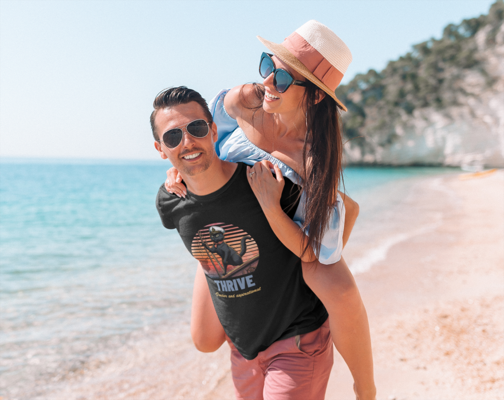 A man is wearing a Black Cat Sunset: A Voyage to THRIVE T-Shirt and is carrying a woman on his back.